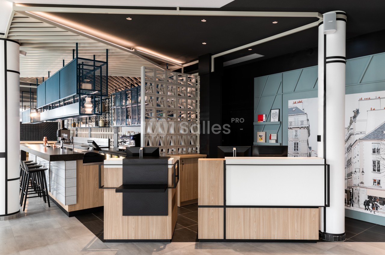 Accueil d'hôtel moderne avec comptoir de réception en bois clair et noir, bar à gauche avec tabourets, et une grande fresque de façades parisiennes sur le mur de droite.