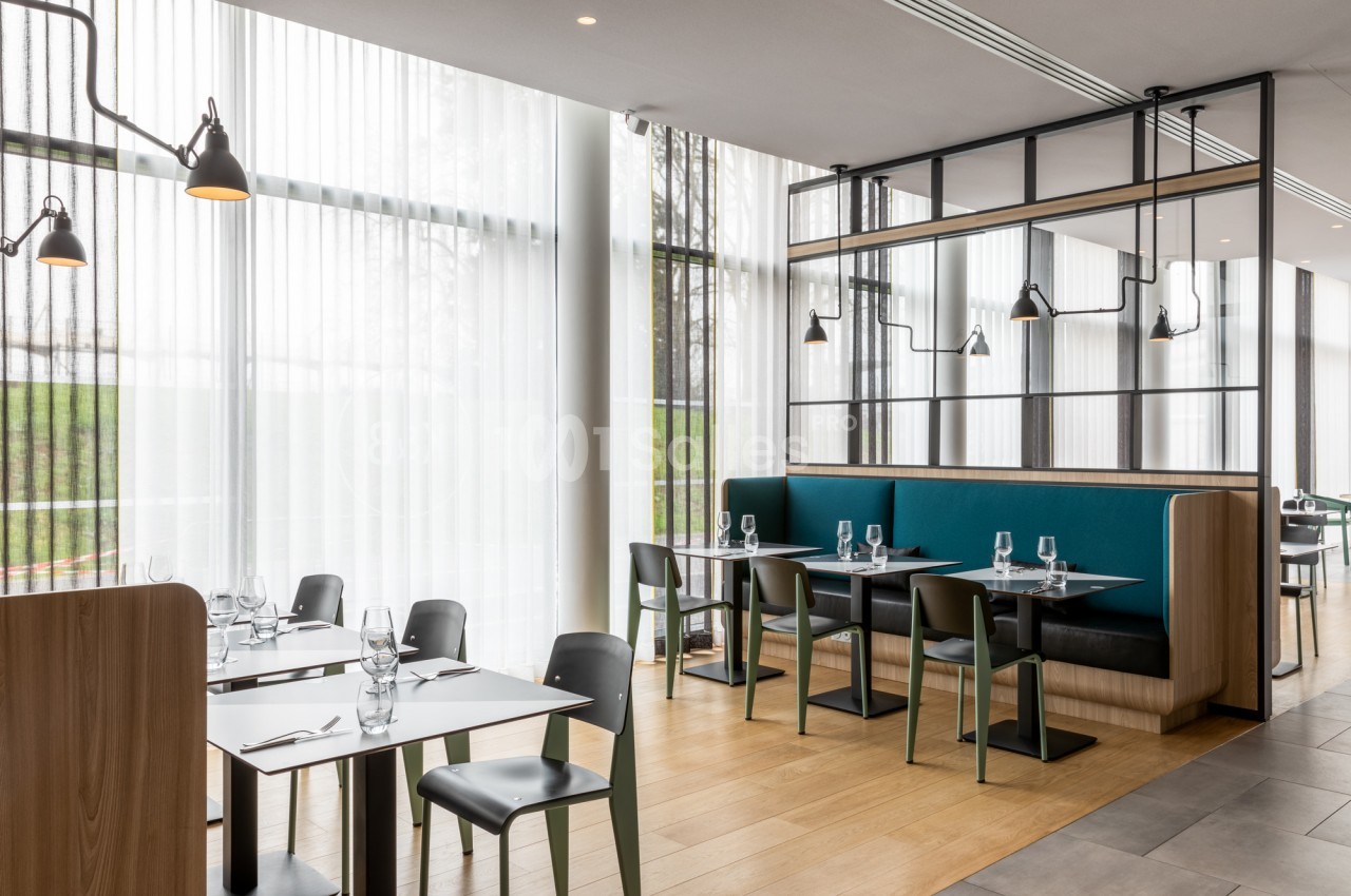Salle de restaurant aux tables carrées dressées de verres et couverts, avec banquettes bleu canard, chaises noires et vertes et grandes baies vitrées voilées de rideaux blancs.