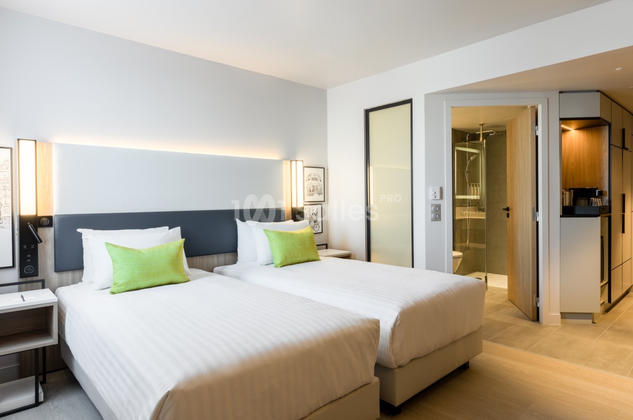 Chambre d'hôtel avec deux lits simples recouverts de linge blanc, coussins verts, tête de lit sombre éclairée et salle de bains visible à droite. Let me check length: roughly 125 characters? Count: "Chambre d'hôtel avec deux lits simples recouverts de