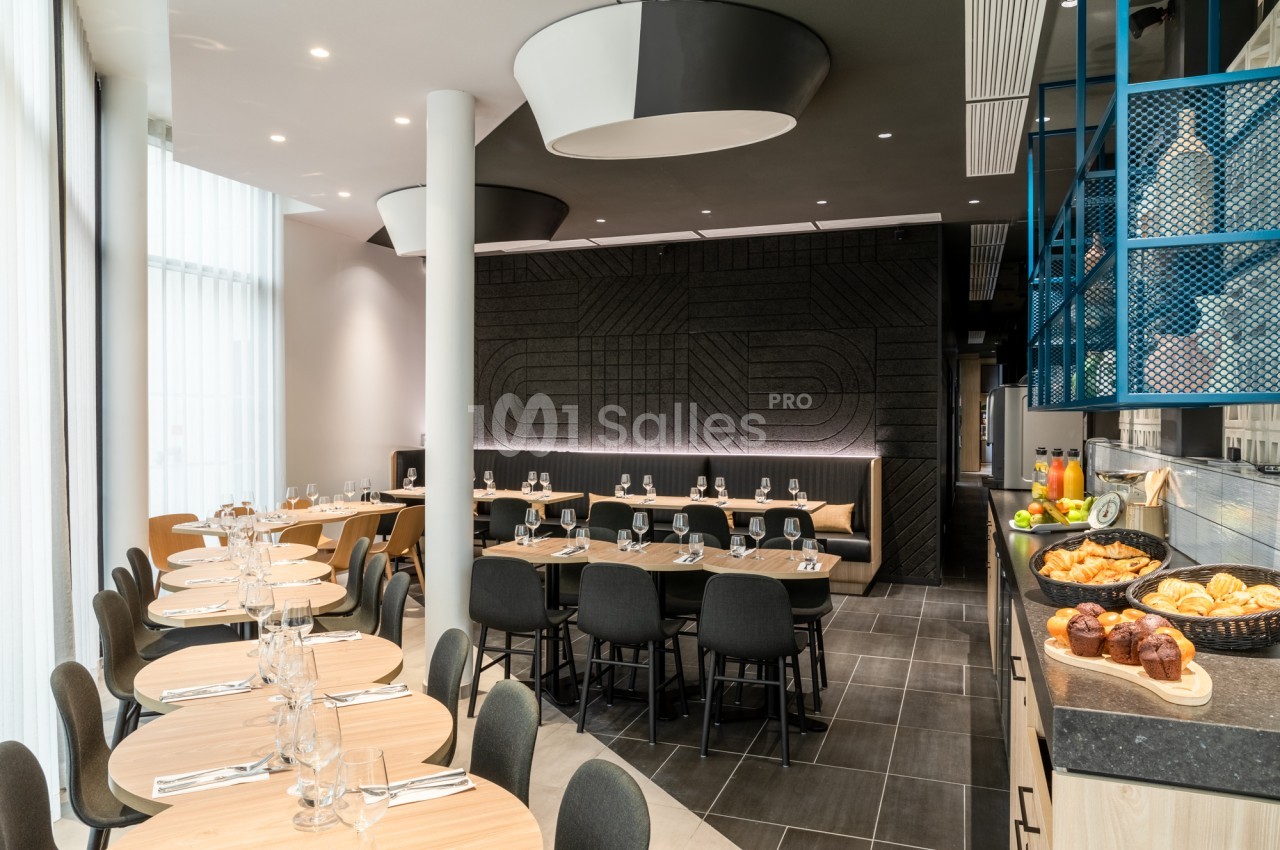 Salle de restaurant aux tables rondes en bois dressées de verres et couverts, chaises noires, et buffet de viennoiseries à droite.