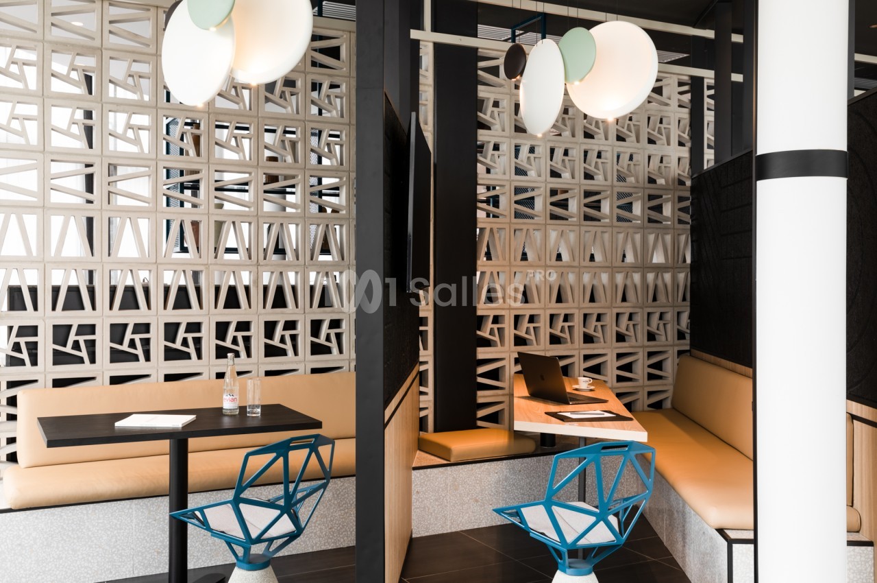 Espace de convivialité avec banquettes en cuir clair, tables noires, chaises bleues design et murs en claustras ajourés, éclairé par des suspensions blanches.