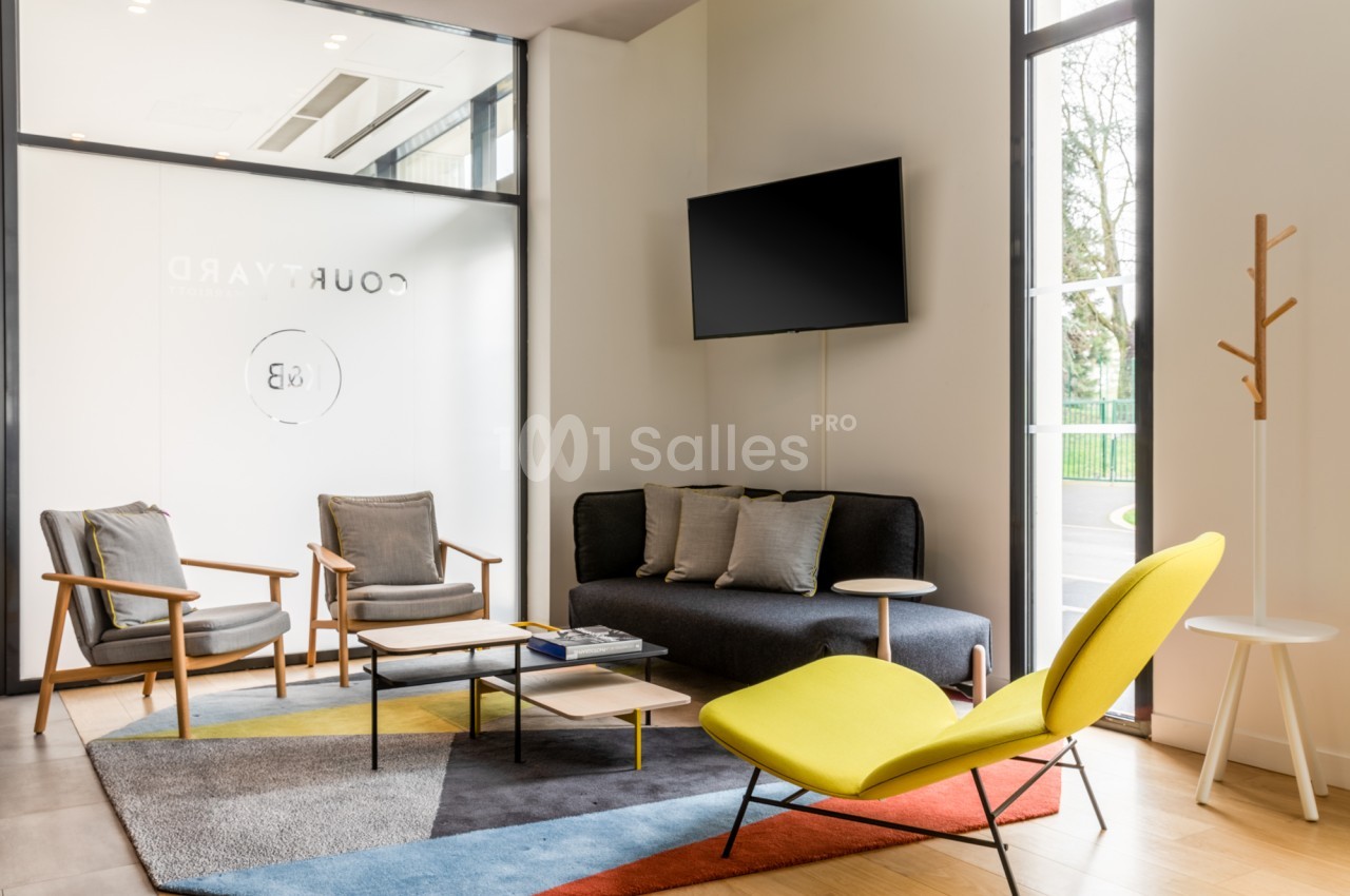Salon d'accueil lumineux avec deux fauteuils gris, un canapé anthracite, un fauteuil jaune, un tapis multicolore et un téléviseur mural.