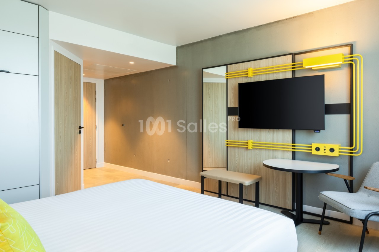 Chambre d'hôtel moderne avec un grand lit blanc au premier plan, un téléviseur fixé au mur sur un panneau bois et jaune, une table ronde et une chaise grise. Wait, I should check length: count roughly. "Chambre d'hôtel moderne avec un grand lit blanc au
