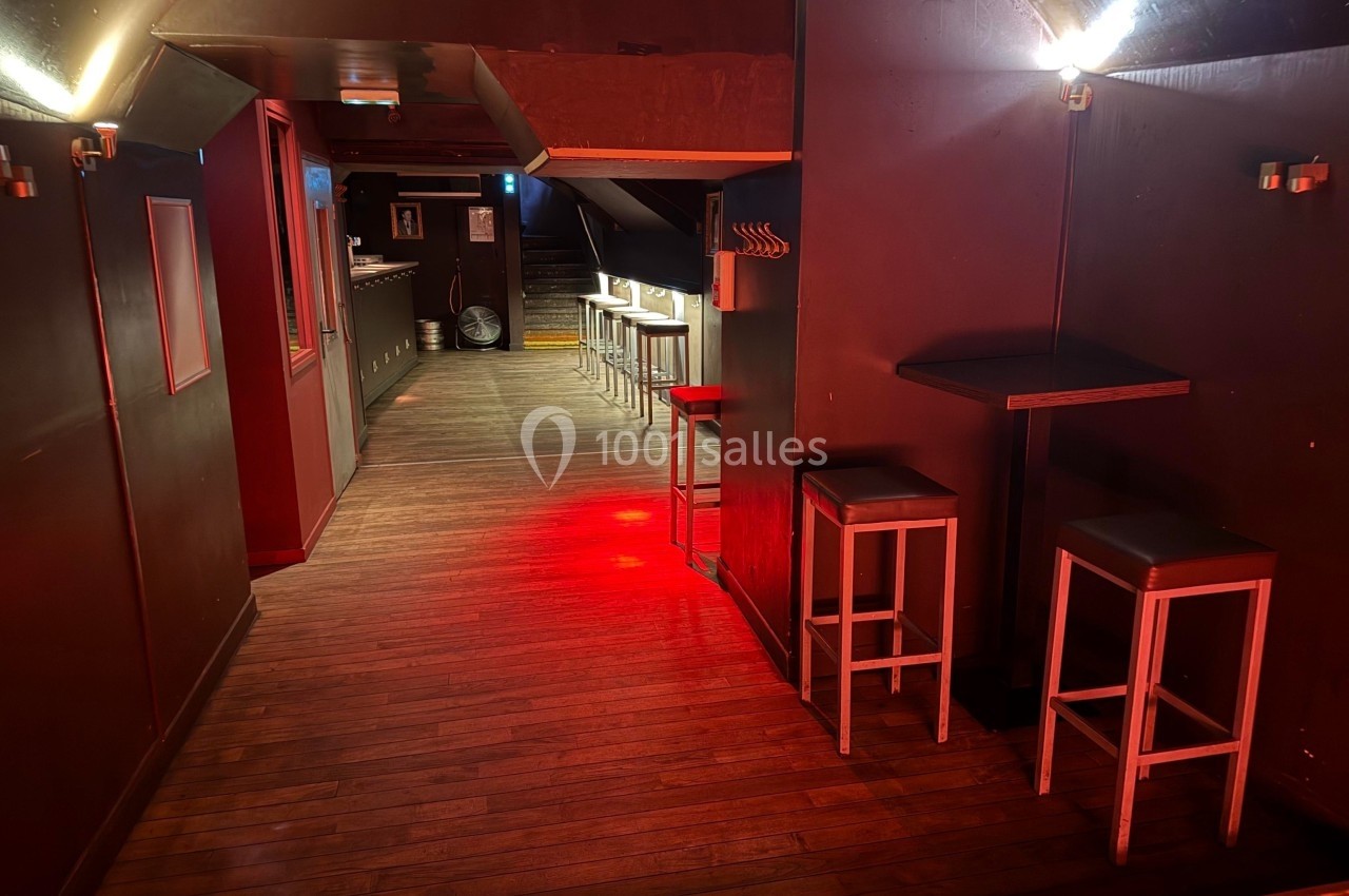 Salle sombre avec parquet en bois, tables hautes et tabourets, éclairage tamisé rouge et comptoir au fond.