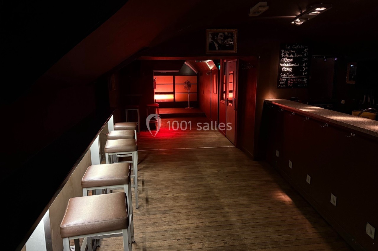 Salle sombre avec un éclairage rouge au fond, tabourets alignés à gauche et un comptoir à droite.