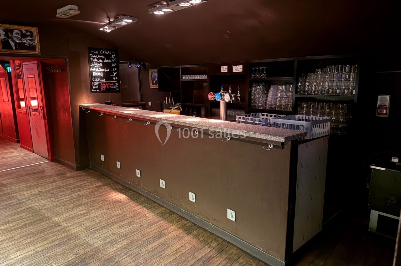 Bar en bois sombre avec comptoir équipé de crochets, étagères à verres et tableau noir affichant le menu.