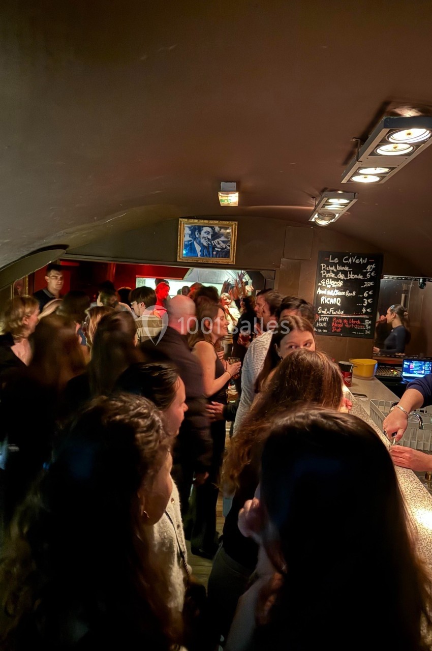 Groupe de personnes discutant et buvant dans un bar animé avec un comptoir éclairé et une ardoise au mur.