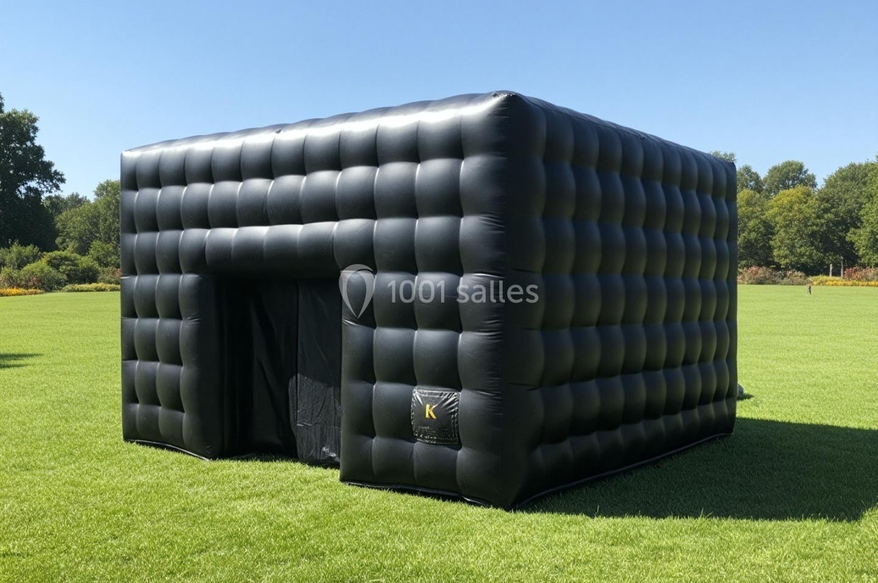 Structure gonflable noire en forme de cube, installée sur une pelouse verte par temps ensoleillé.