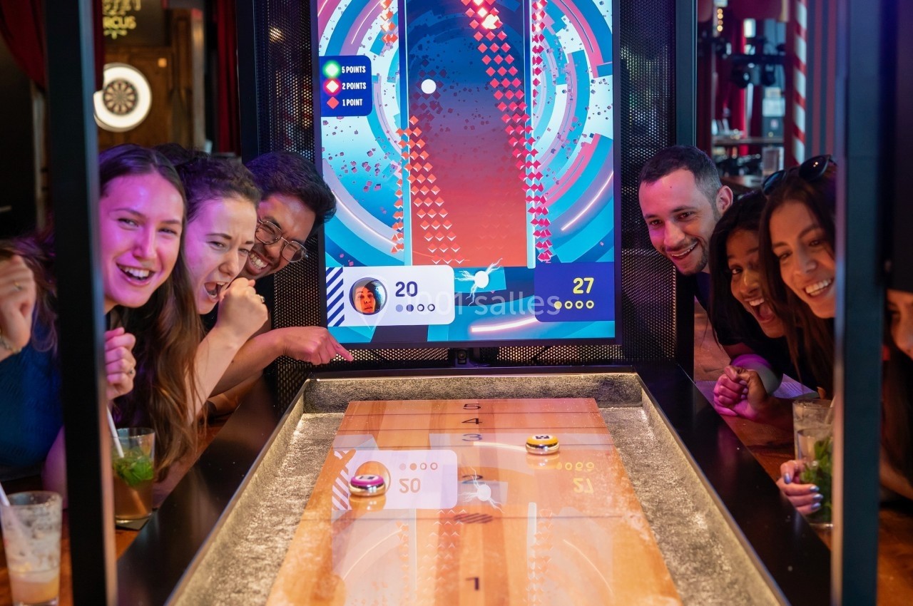 Des personnes jouent à un jeu de palets interactif dans un bar, avec un écran affichant les scores en arrière-plan.