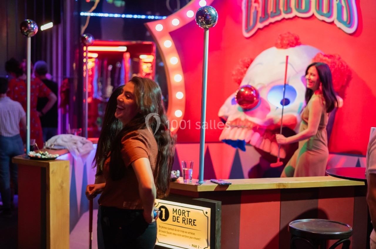 Deux femmes souriantes dans un décor de fête foraine avec un stand coloré et une tête de clown en arrière-plan.