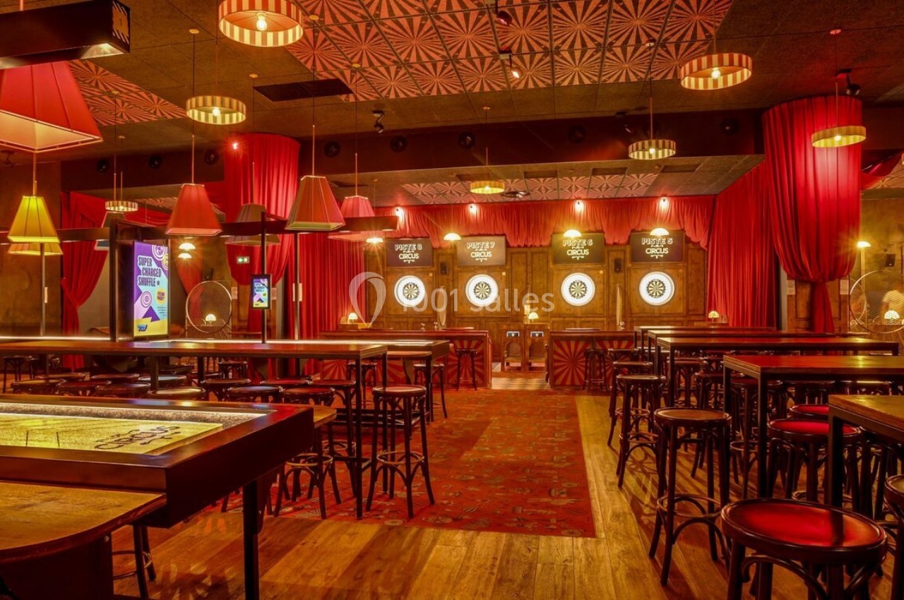 Salle de jeux avec cibles de fléchettes éclairées, tables hautes en bois et rideaux rouges en arrière-plan.