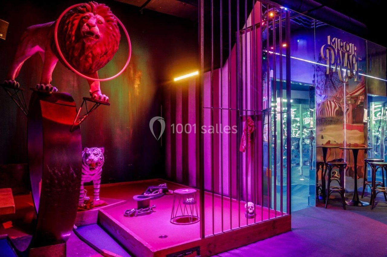 Décor de cirque avec un lion sur un socle, un tigre au sol et une cage éclairée par des lumières colorées.