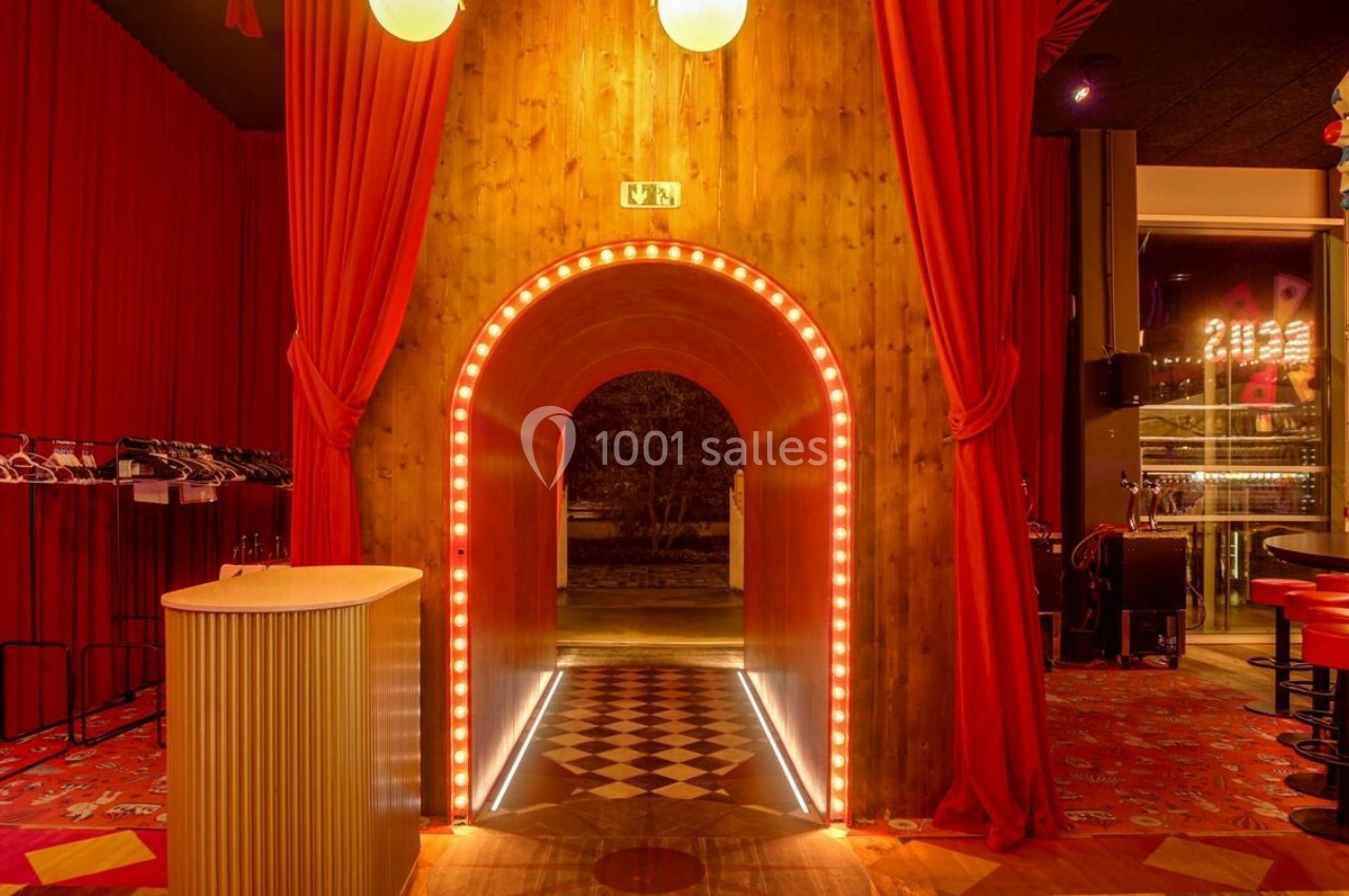 Entrée voûtée éclairée par des lumières, entourée de rideaux rouges, avec un sol à motifs géométriques.