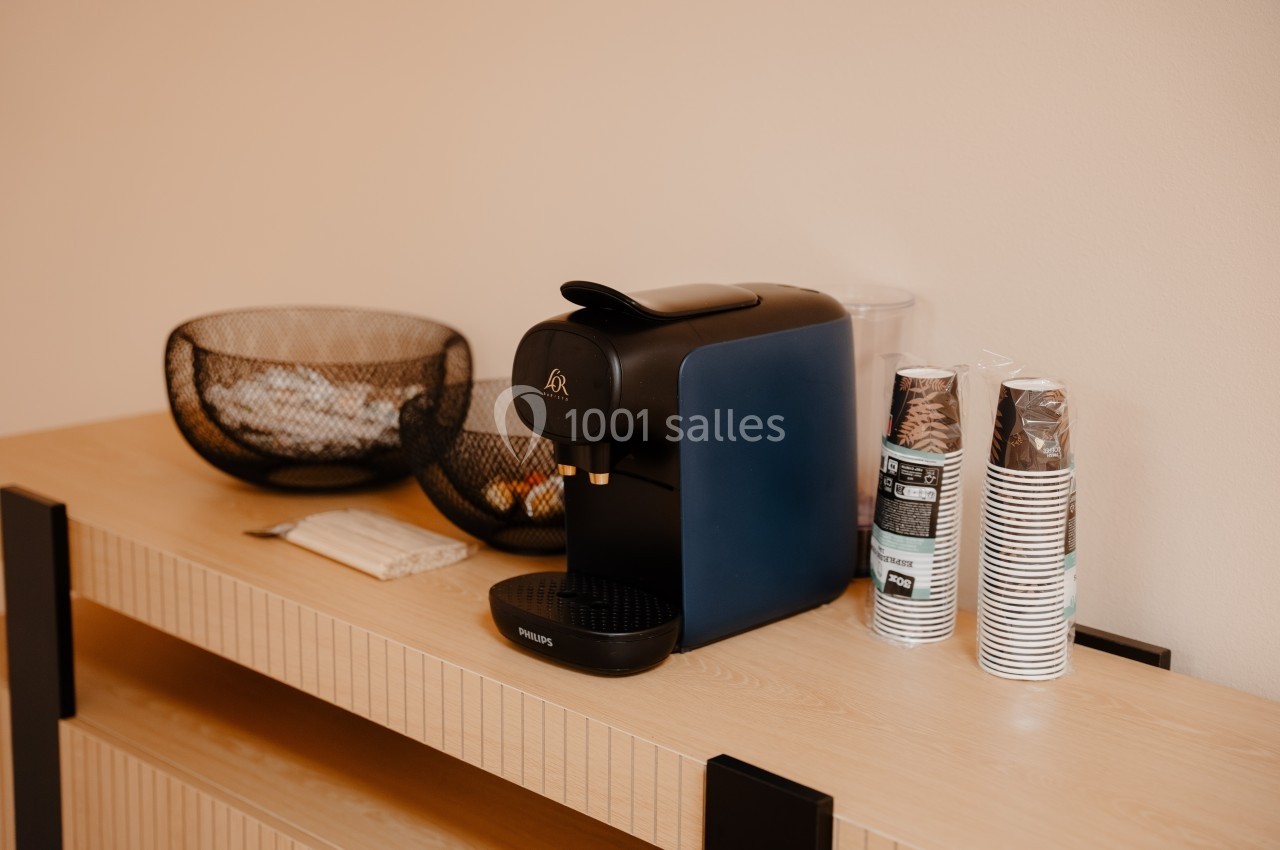 Machine à café noire sur un meuble en bois clair, entourée de gobelets en carton et de paniers en métal.