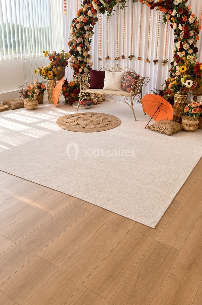 Décoration florale avec arche de fleurs colorées, banc en métal, tapis rond et parasols orange dans une pièce lumineuse.