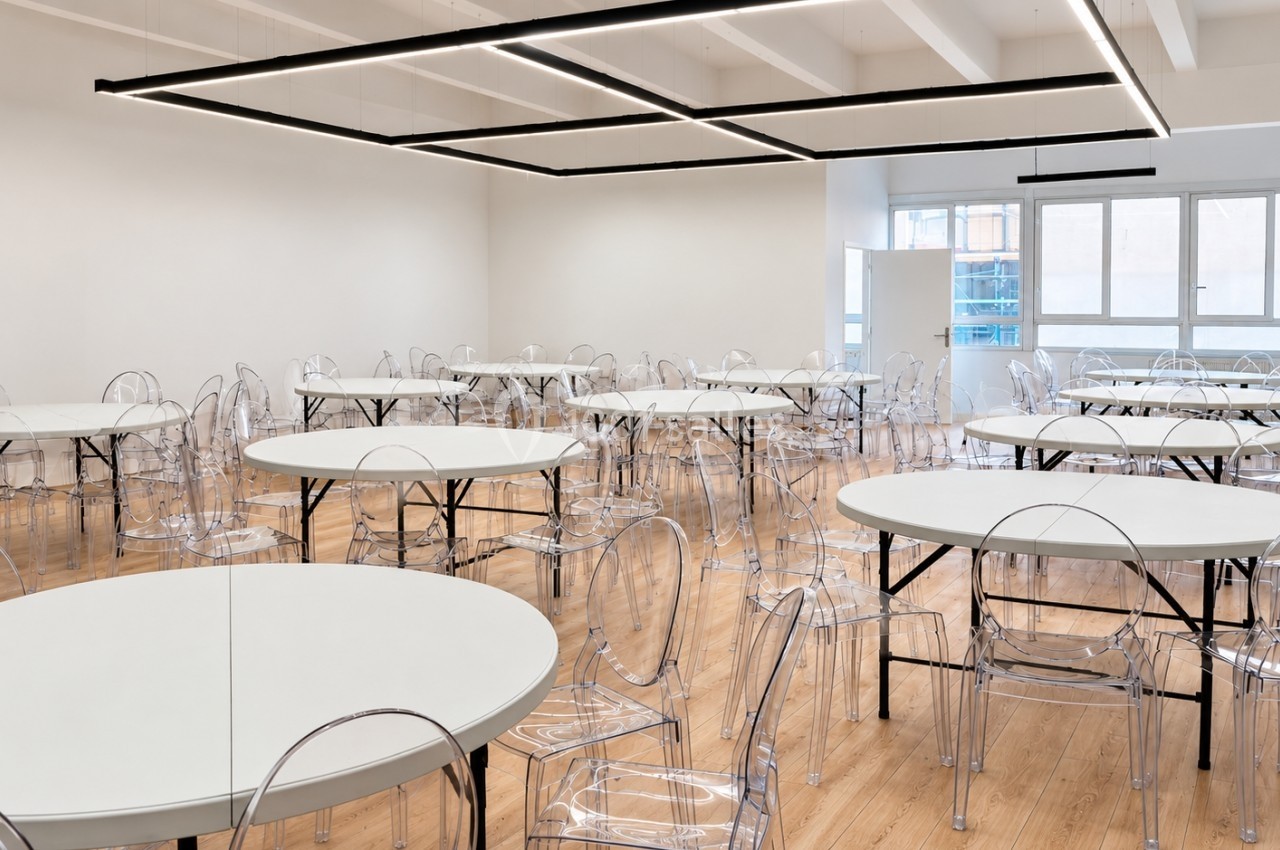 Salle lumineuse avec tables rondes blanches et chaises transparentes disposées sur un sol en bois clair.