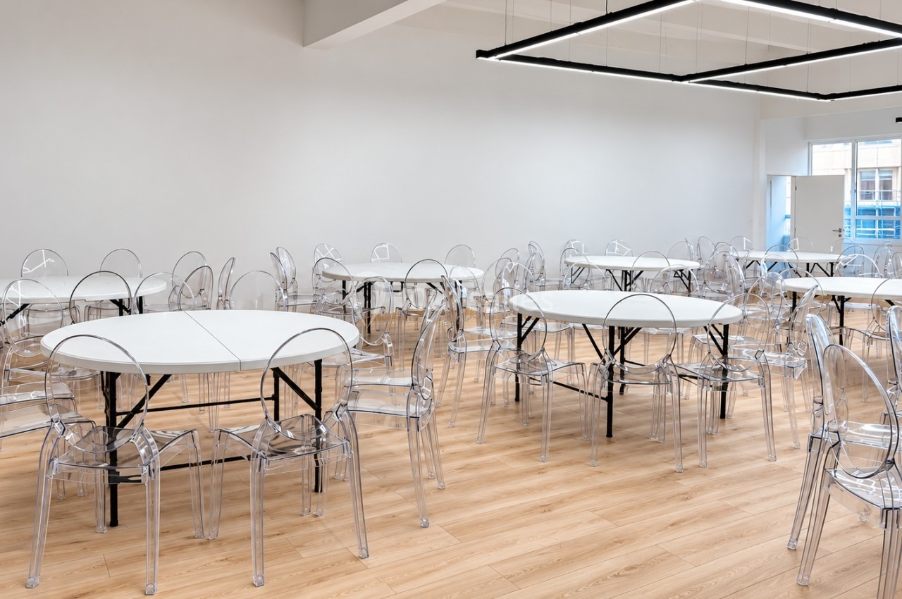 Salle lumineuse avec des tables rondes blanches et des chaises transparentes sur un sol en bois clair.