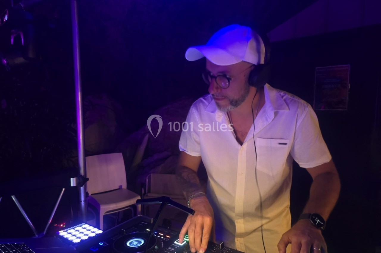 Un DJ en chemise blanche et casquette mixe de la musique sur une table de mixage dans une ambiance nocturne.