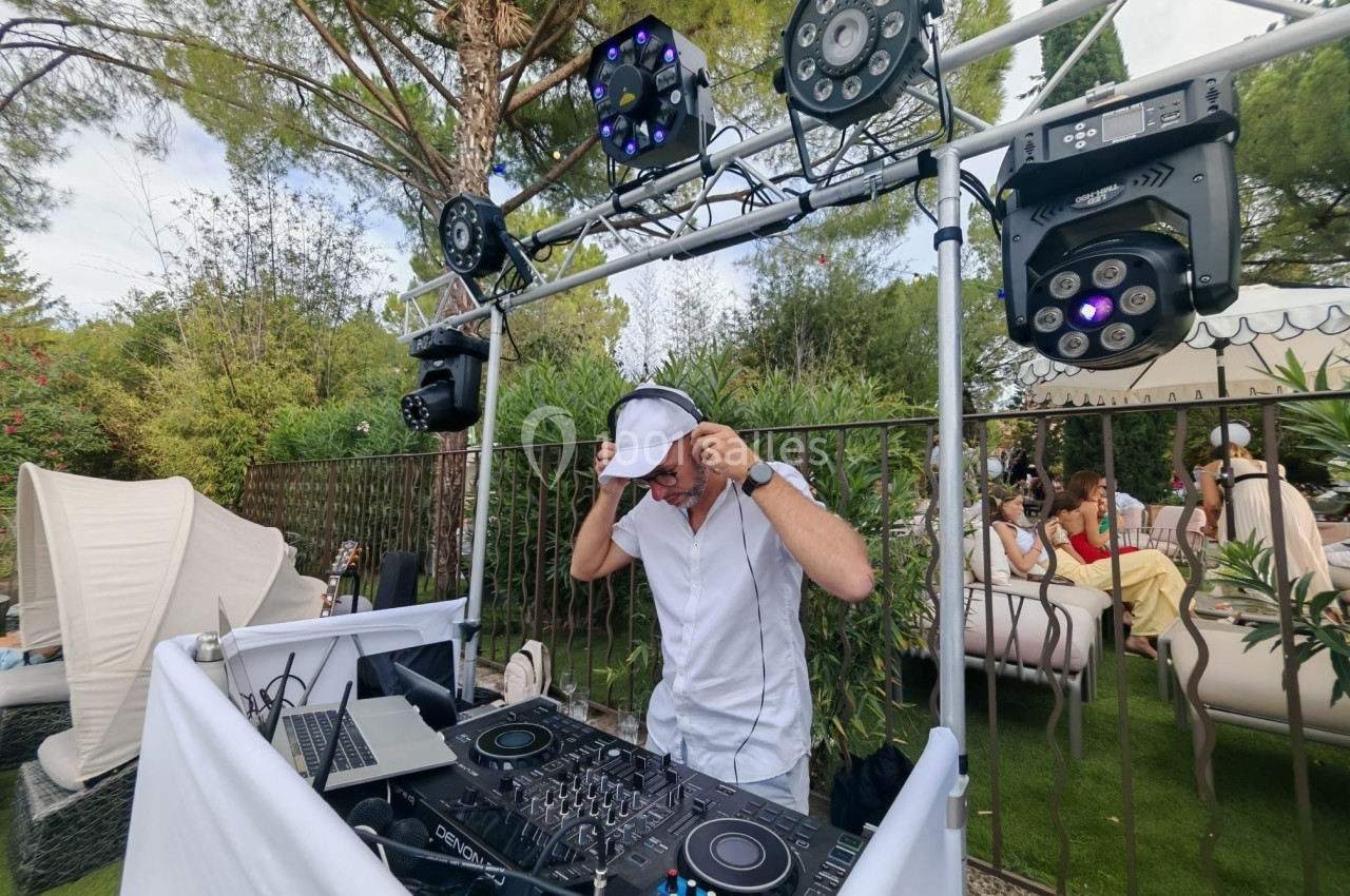 Un DJ mixe en extérieur sous un éclairage scénique, entouré de végétation et de personnes assises en arrière-plan.
