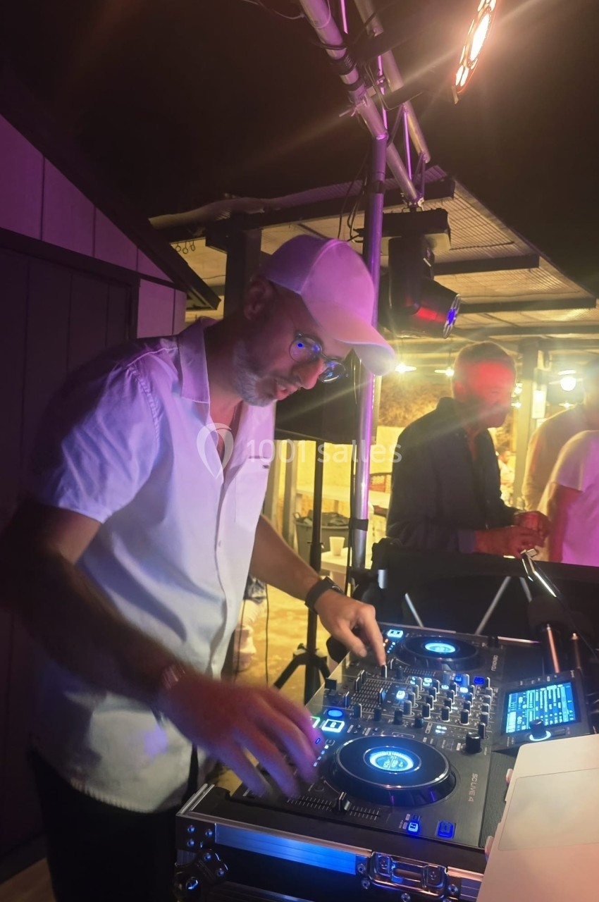 Un DJ mixe de la musique sur une table de mixage éclairée, dans une ambiance nocturne avec des lumières colorées.