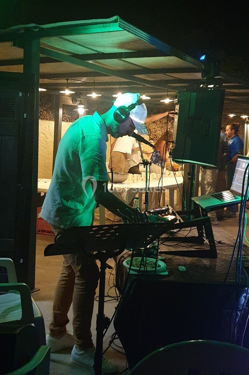 Un DJ mixe de la musique en extérieur, sous un éclairage tamisé, avec des invités en arrière-plan.