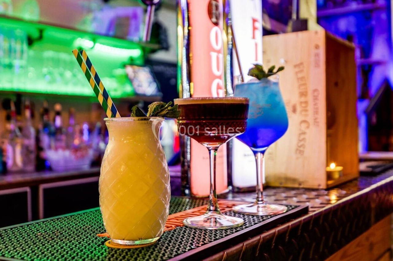 Trois cocktails colorés alignés sur un comptoir de bar, décorés de menthe et entourés d'une ambiance lumineuse.