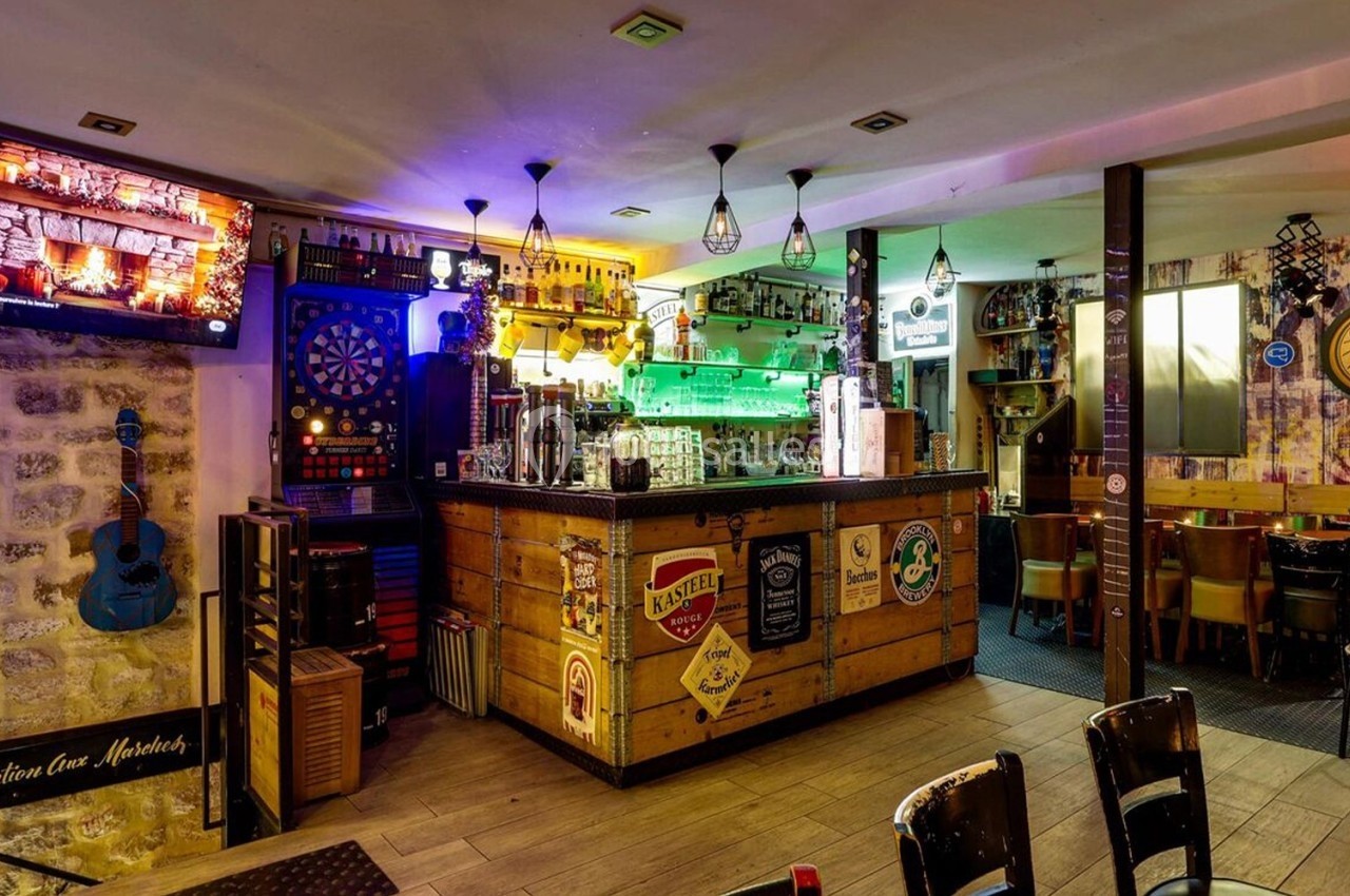 Intérieur d'un bar avec comptoir en bois, décorations variées, écran mural, cible de fléchettes et tables disposées autour.
