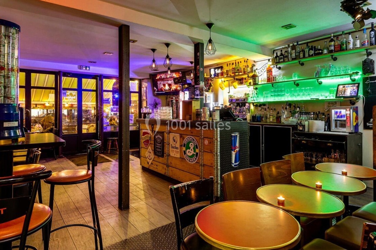 Intérieur d'un bar avec comptoir éclairé, tables rondes, chaises hautes et ambiance lumineuse colorée.