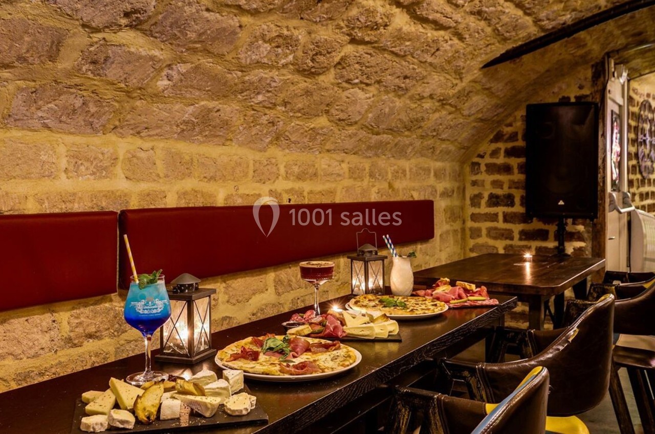 Salle voûtée en pierre avec tables dressées, plats variés, cocktails colorés et ambiance chaleureuse.