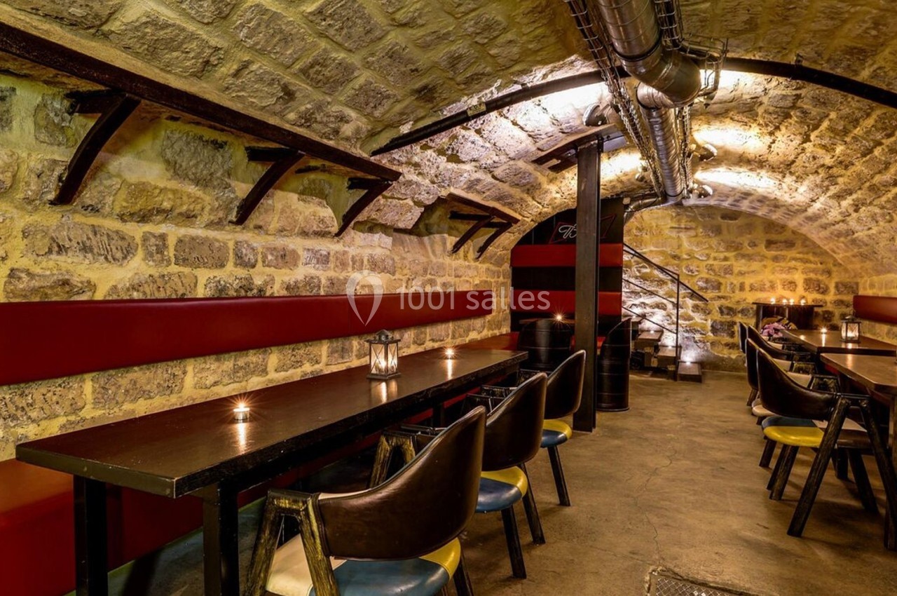 Salle voûtée en pierre avec tables et chaises alignées, éclairage tamisé et escalier en fond.