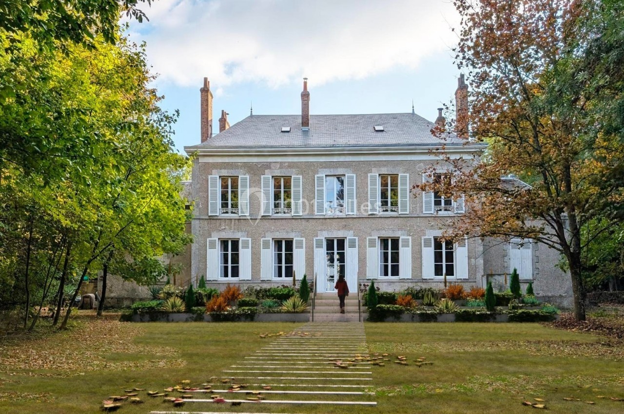 Façade d'une grande maison en pierre entourée d'arbres et d'un jardin avec une allée centrale.