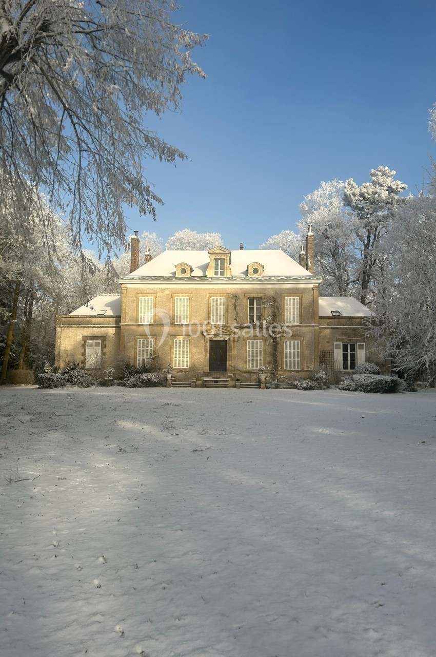 Manoir entouré d'arbres enneigés sous un ciel bleu clair, avec un jardin recouvert de neige.