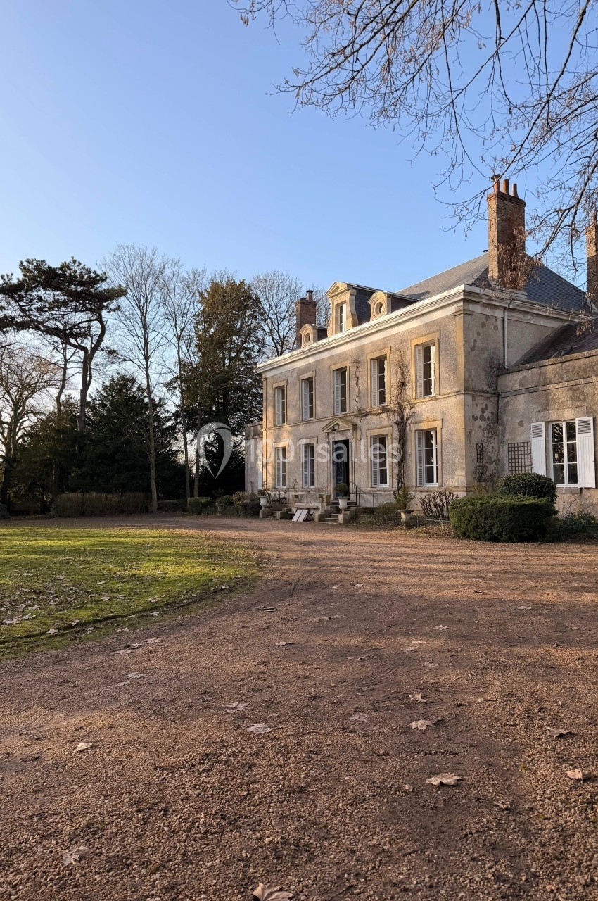 Façade d'un manoir ancien entouré d'arbres, avec une allée en terre au premier plan sous un ciel dégagé.