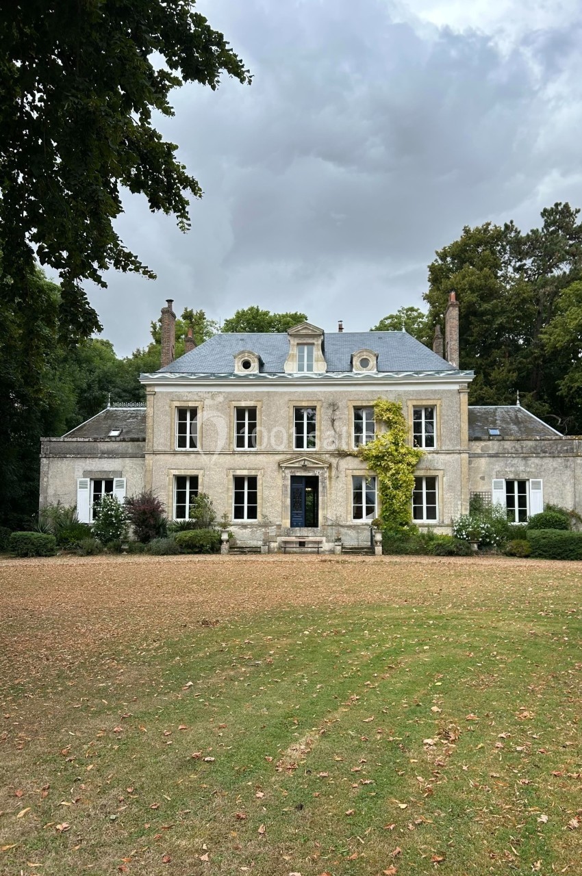 Façade d'une maison de campagne en pierre, entourée d'arbres, avec une pelouse partiellement jaunie au premier plan.