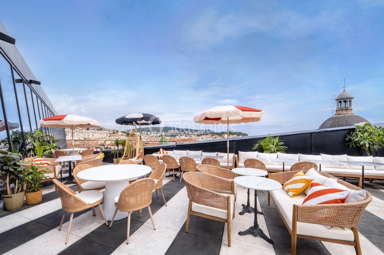 Terrasse en rooftop avec mobilier en rotin, parasols colorés et vue panoramique sur une ville et des collines.