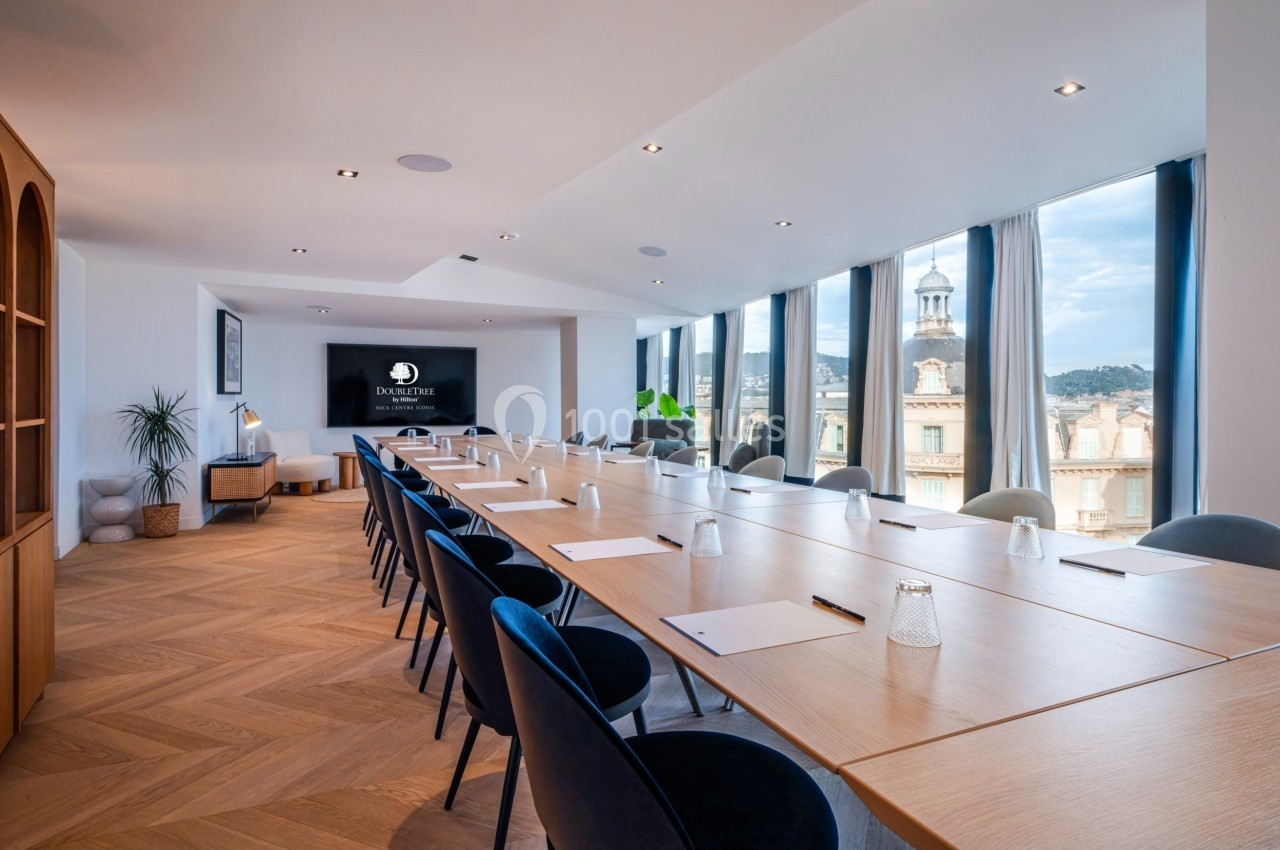 Salle de réunion lumineuse avec une grande table en bois, chaises bleues, écran mural et vue sur des bâtiments extérieurs.