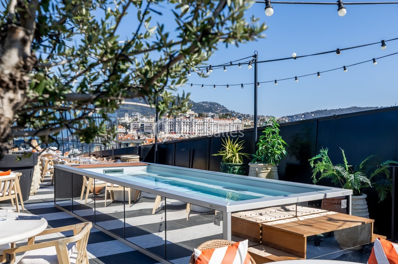 Terrasse avec piscine étroite, mobilier extérieur, plantes et vue sur une ville côtière sous un ciel dégagé.