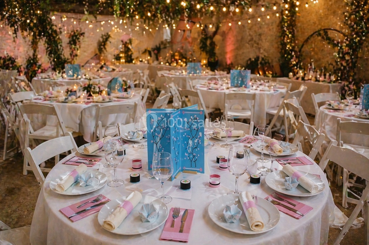 Salle de réception décorée avec des tables rondes dressées, ornées de menus bleus et éclairées par des guirlandes lumineuses.