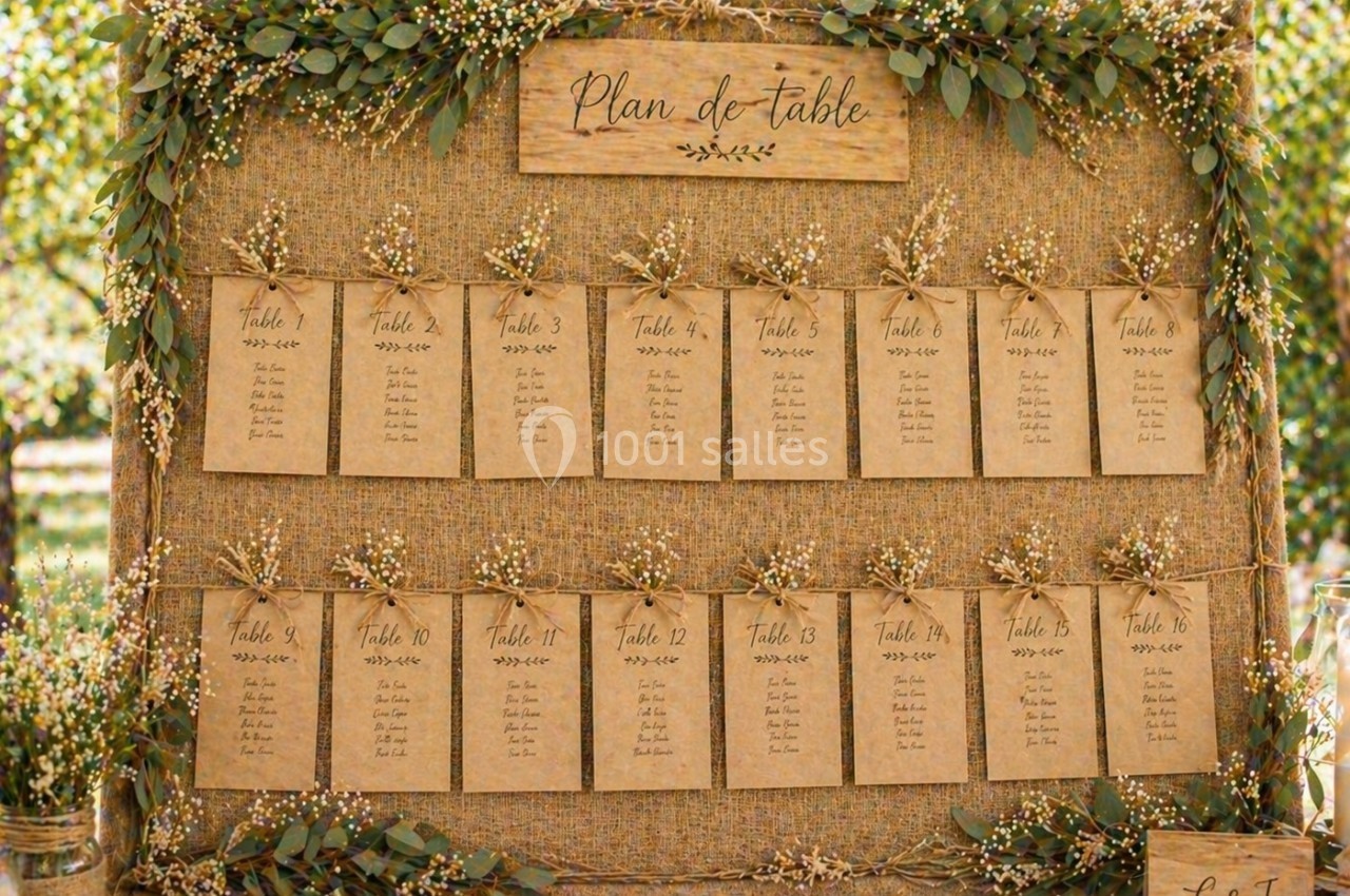 Plan de table rustique avec cartes en papier kraft suspendues sur une toile de jute, décoré de fleurs blanches et feuillages.