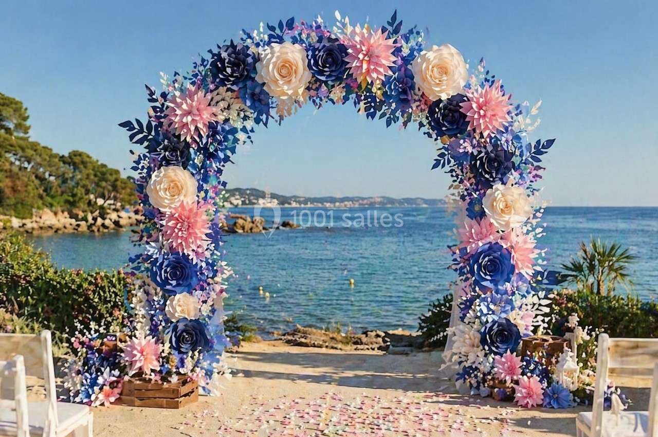 Arche florale décorative avec des fleurs roses et bleues, installée sur une plage avec vue sur la mer et le ciel dégagé.