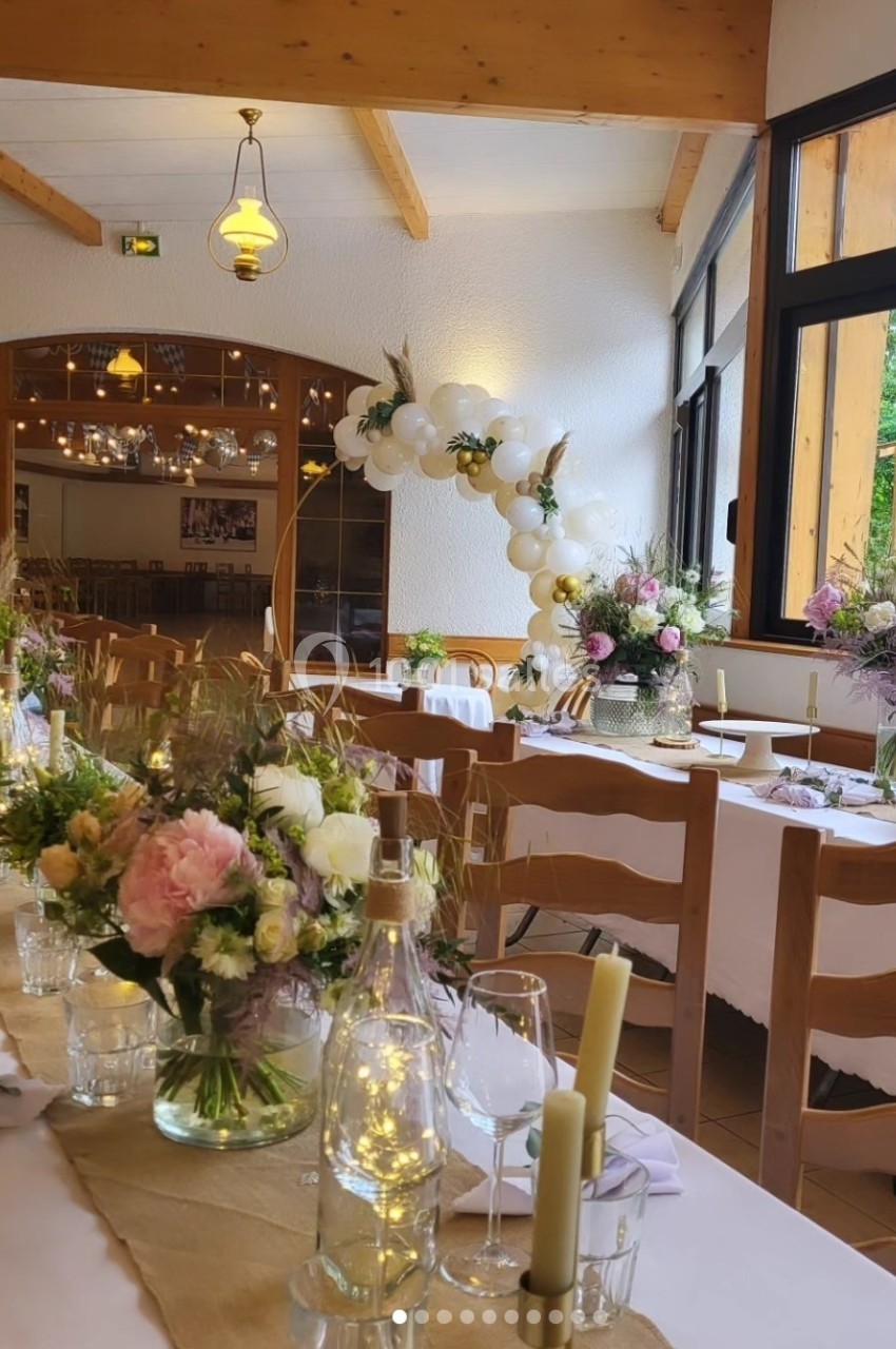 Salle de réception décorée avec des tables dressées, des fleurs pastel et des ballons blancs dans un cadre lumineux.