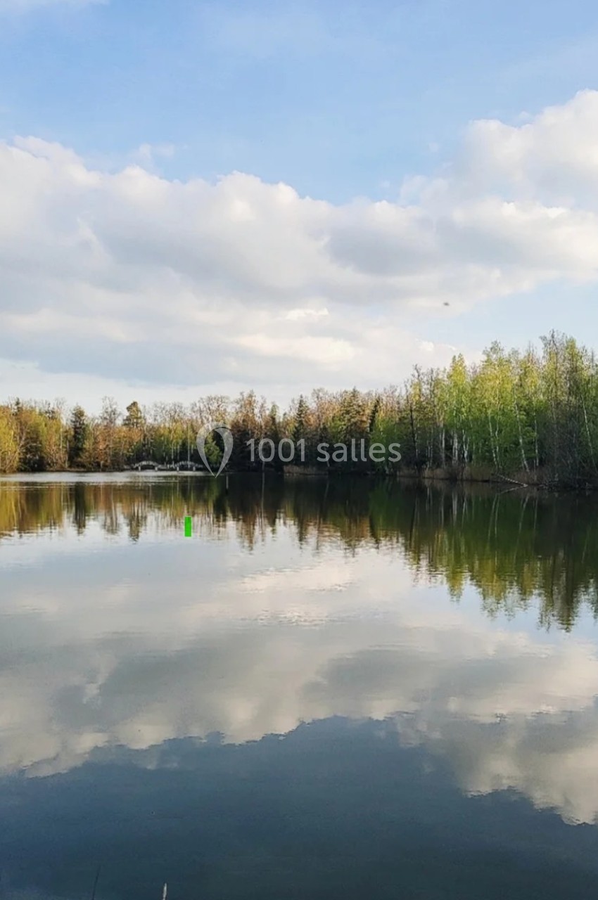 Vue d'un lac calme reflétant des arbres et un ciel partiellement nuageux, entouré de végétation.