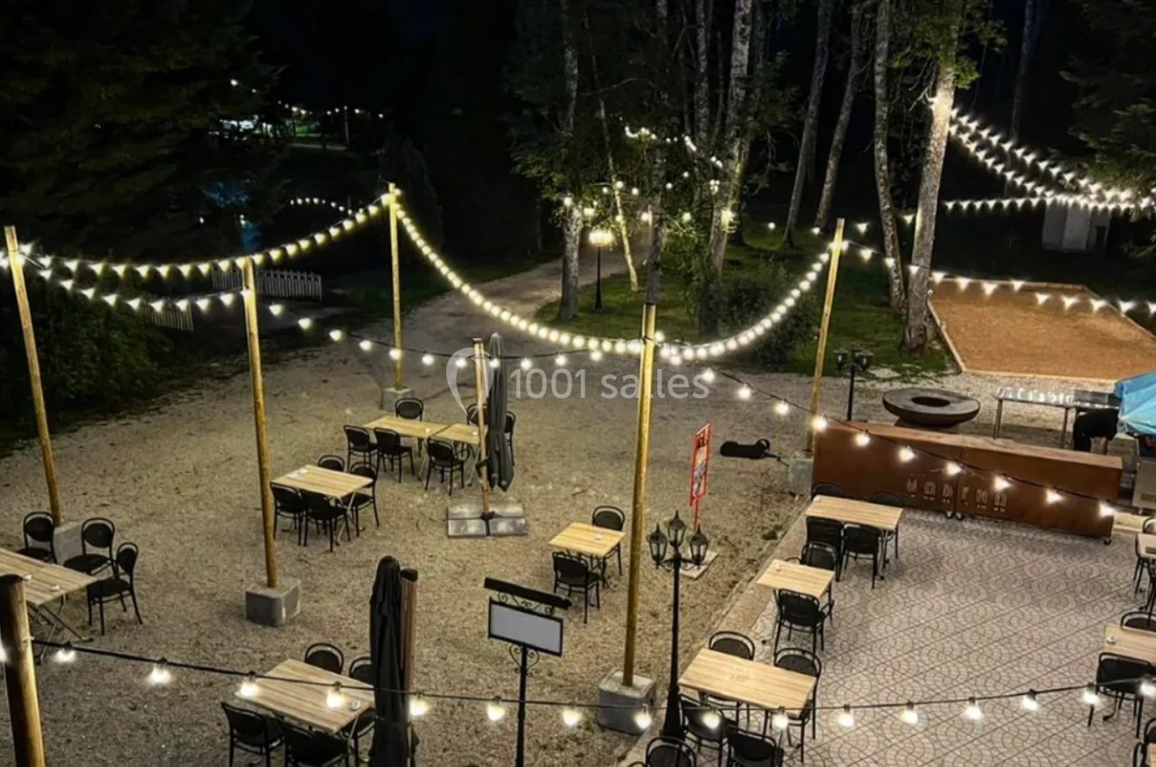 Terrasse extérieure éclairée par des guirlandes lumineuses, avec tables et chaises disposées sur un sol en gravier.