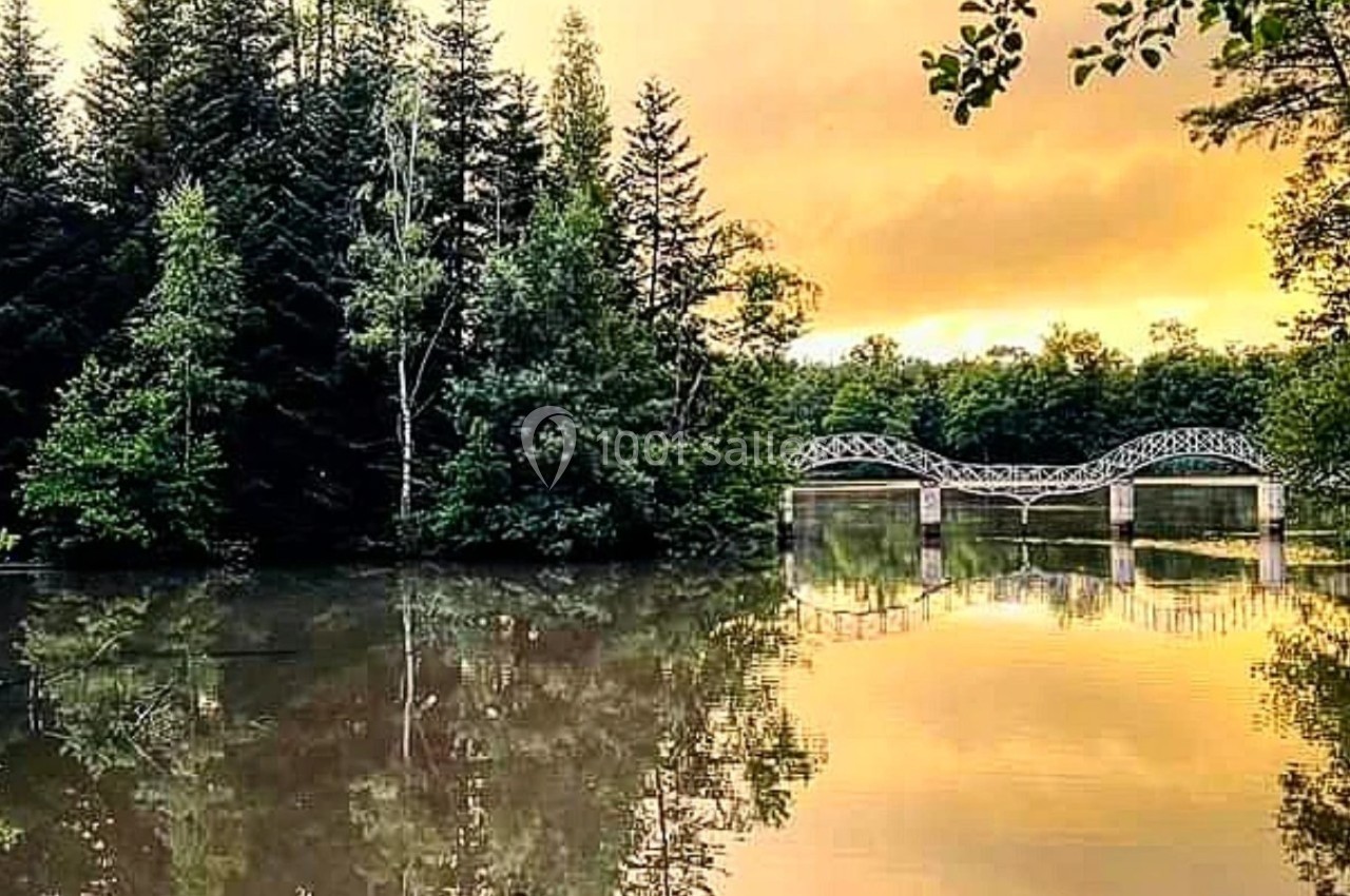 Pont en arc blanc traversant une rivière calme entourée de forêt, sous un ciel orange au coucher du soleil.