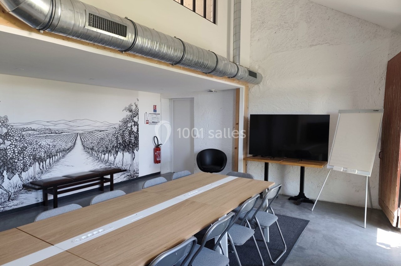 Salle de réunion lumineuse avec table en bois, chaises pliantes, écran, paperboard et fresque murale représentant des vignes.