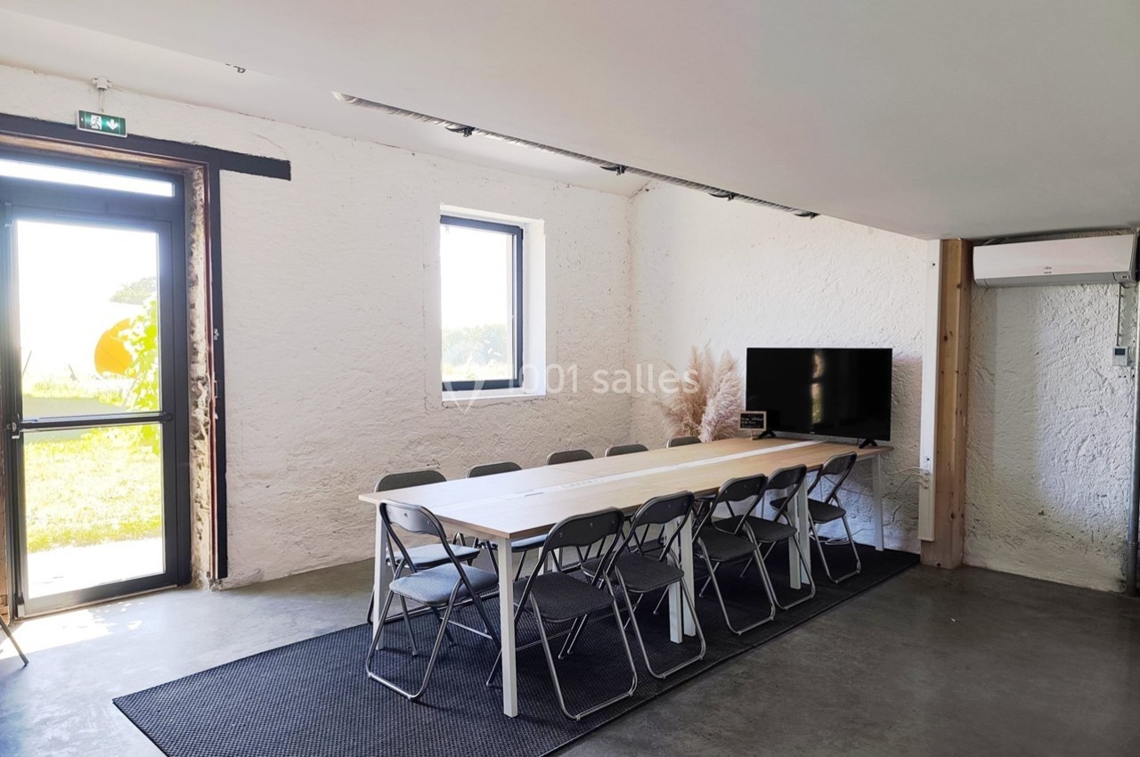 Salle lumineuse avec une grande table en bois entourée de chaises pliantes, un écran et une porte donnant sur l'extérieur.