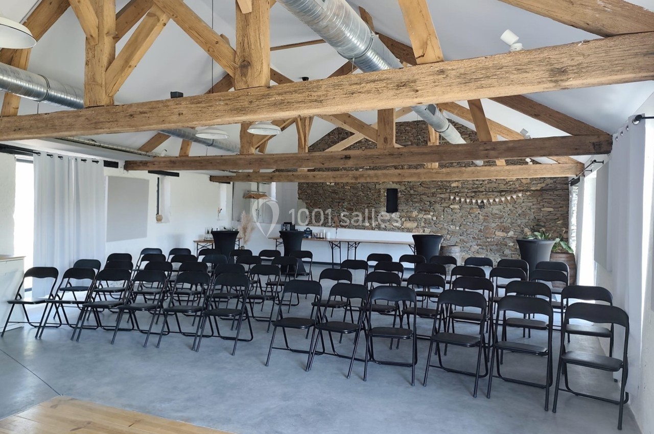 Salle lumineuse avec poutres apparentes, chaises noires alignées face à un espace avec comptoir et murs en pierre.