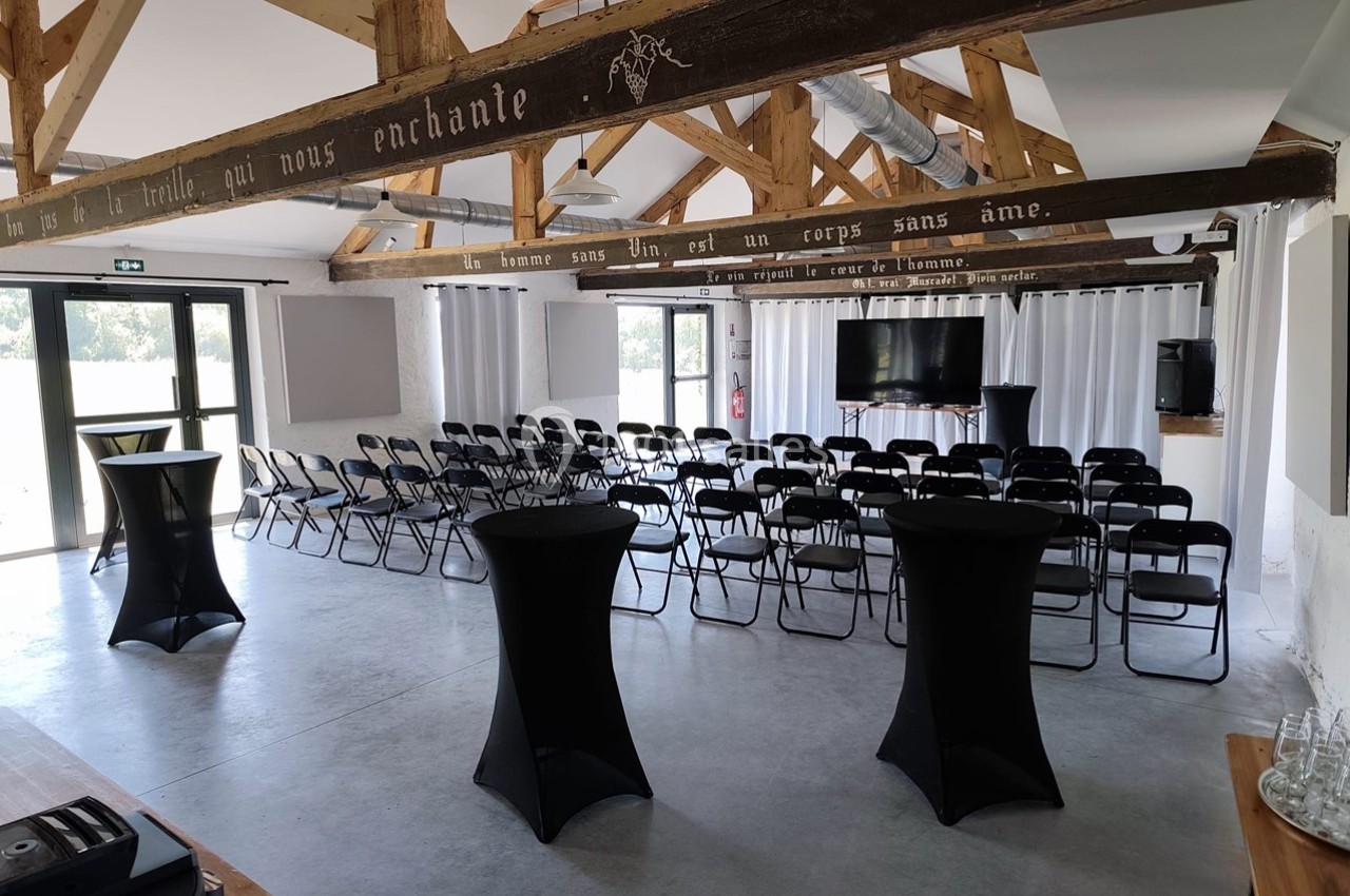 Salle de conférence lumineuse avec des rangées de chaises noires, tables hautes et poutres apparentes en bois.