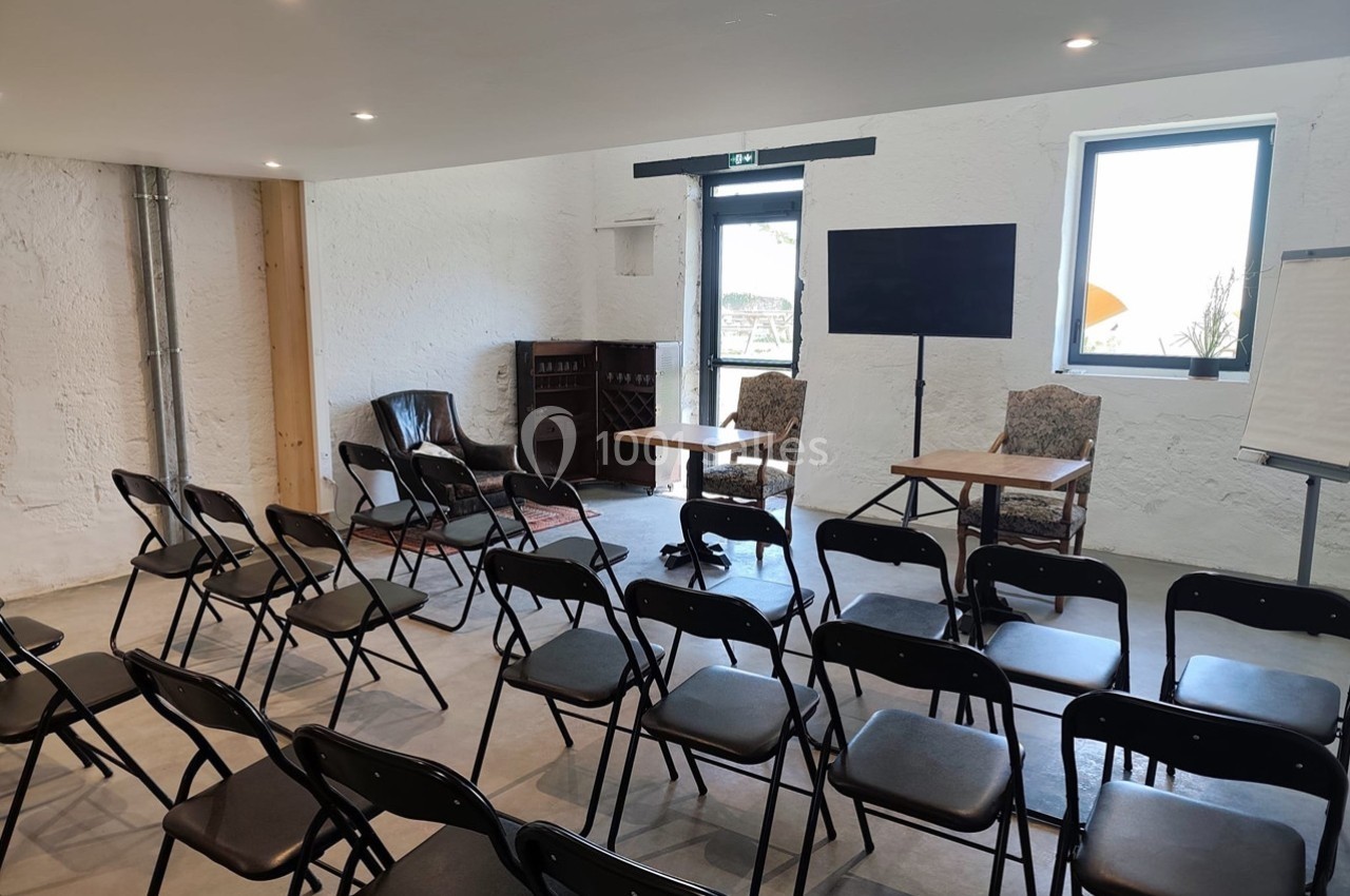Salle de réunion lumineuse avec chaises noires disposées face à deux tables, un écran et un tableau blanc.