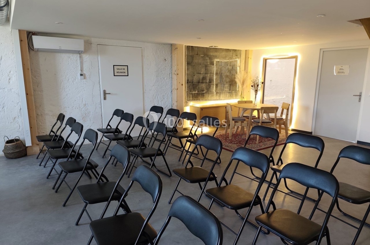 Salle lumineuse avec des chaises pliantes noires disposées en rangées face à une table et un miroir éclairé.